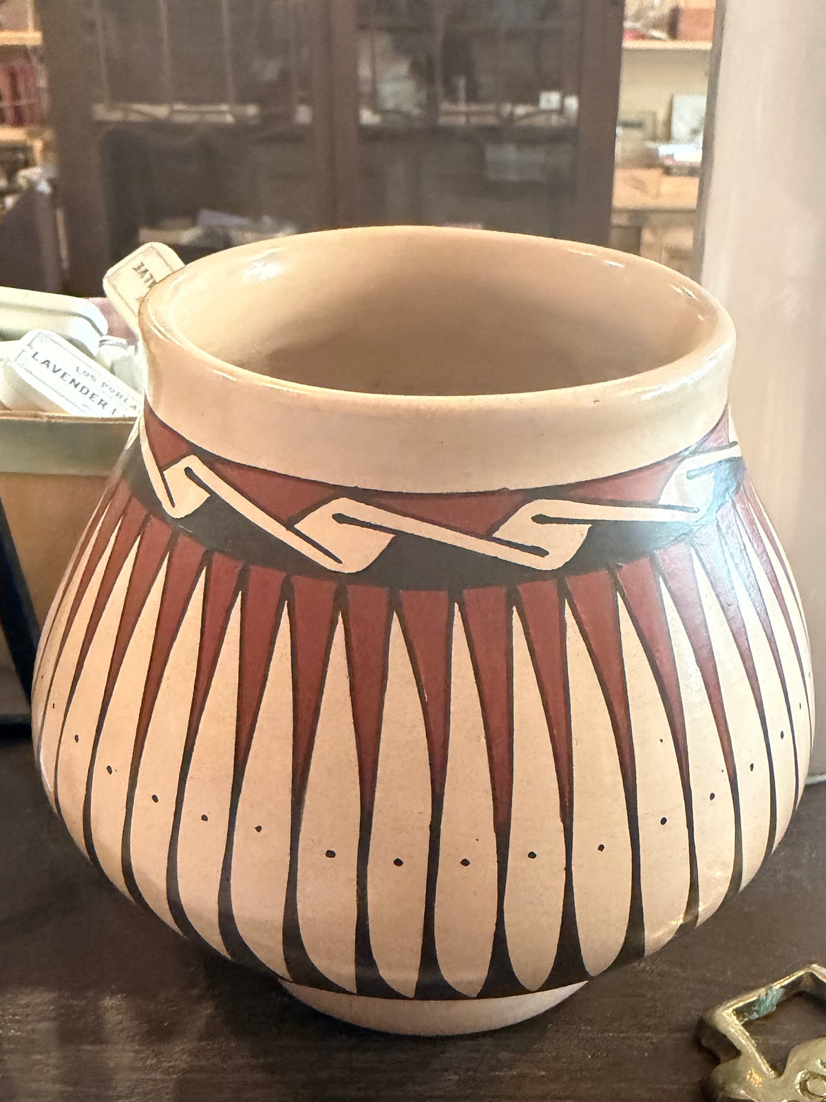 A handmade ceramic pot with a Southwestern geometric pattern, featuring a cream-colored body, brown and black painted stripes, and a beige rim, sitting on a wooden table next to a Sharpie marker.