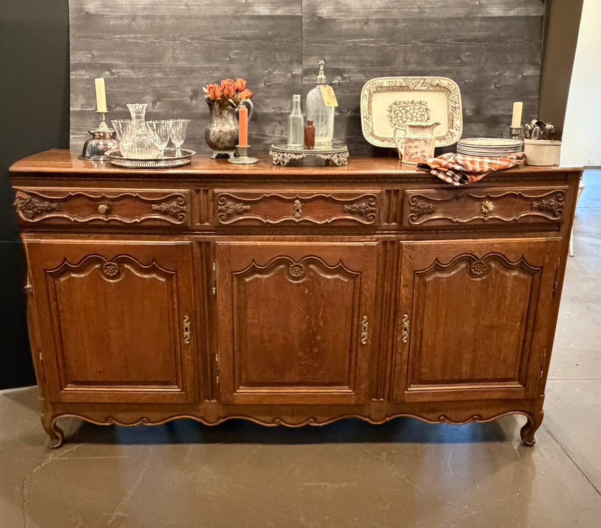 An antique carved wooden sideboard or buffet table with three doors and three drawers, styled with glassware, bottles, and decorative plates on top, set against a dark wood plank wall.