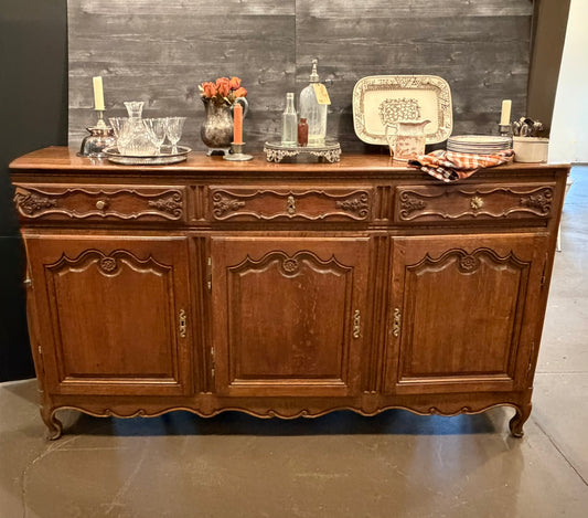 An antique carved wooden sideboard or buffet table with three doors and three drawers, styled with glassware, bottles, and decorative plates on top, set against a dark wood plank wall.