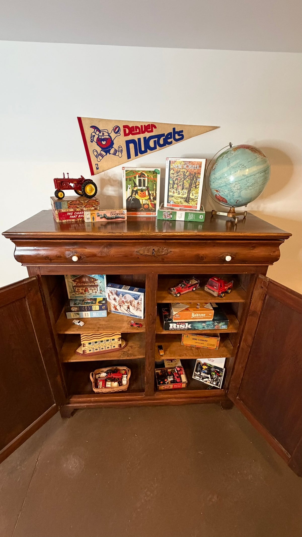 A dark brown wooden toy cabinet with open doors, filled with various vintage toy cars, board games, and collectibles. On top of the cabinet are a red tractor, a 'Dealer Nuggets' sports pennant, a globe, and a small toy car, all set against a plain white wall.