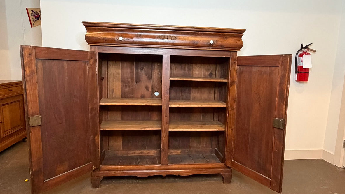 A large rustic wooden bookcase or cabinet with two doors swung open, showing three open shelves inside. The piece of furniture is made of medium-to-dark brown wood with a distressed finish and brass-toned hardware.