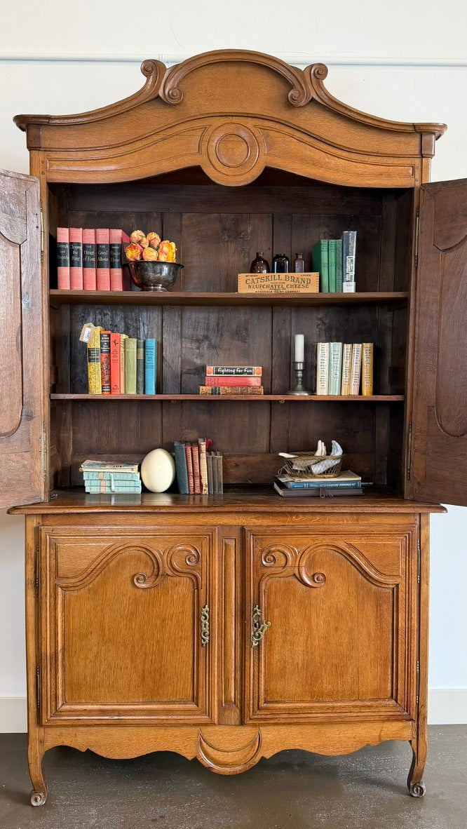 An antique wooden hutch or bookcase with open doors, featuring carved details, five shelves filled with books and decorative items, and two cabinet doors below, photographed against a plain white wall.