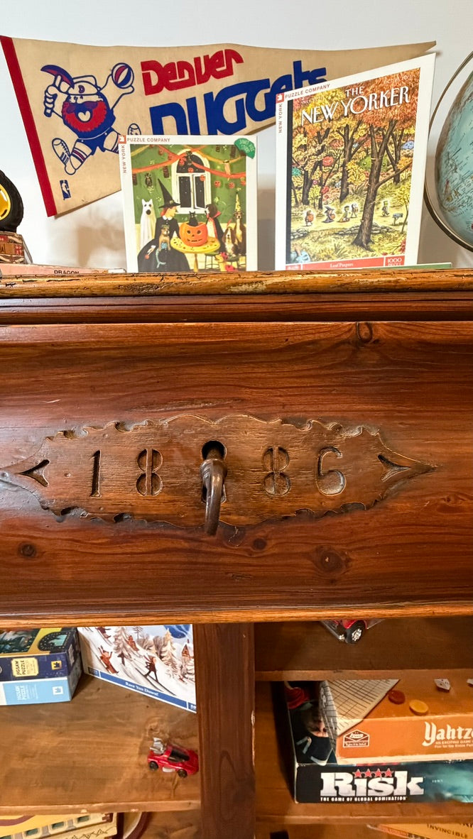 A close-up of a vintage wooden chest of drawers with a carved '1886' handle and a metal pull. Decorative items including a 'Denver Nuggets' banner, a 'New Yorker' magazine, and board games are visible on and around the shelf.