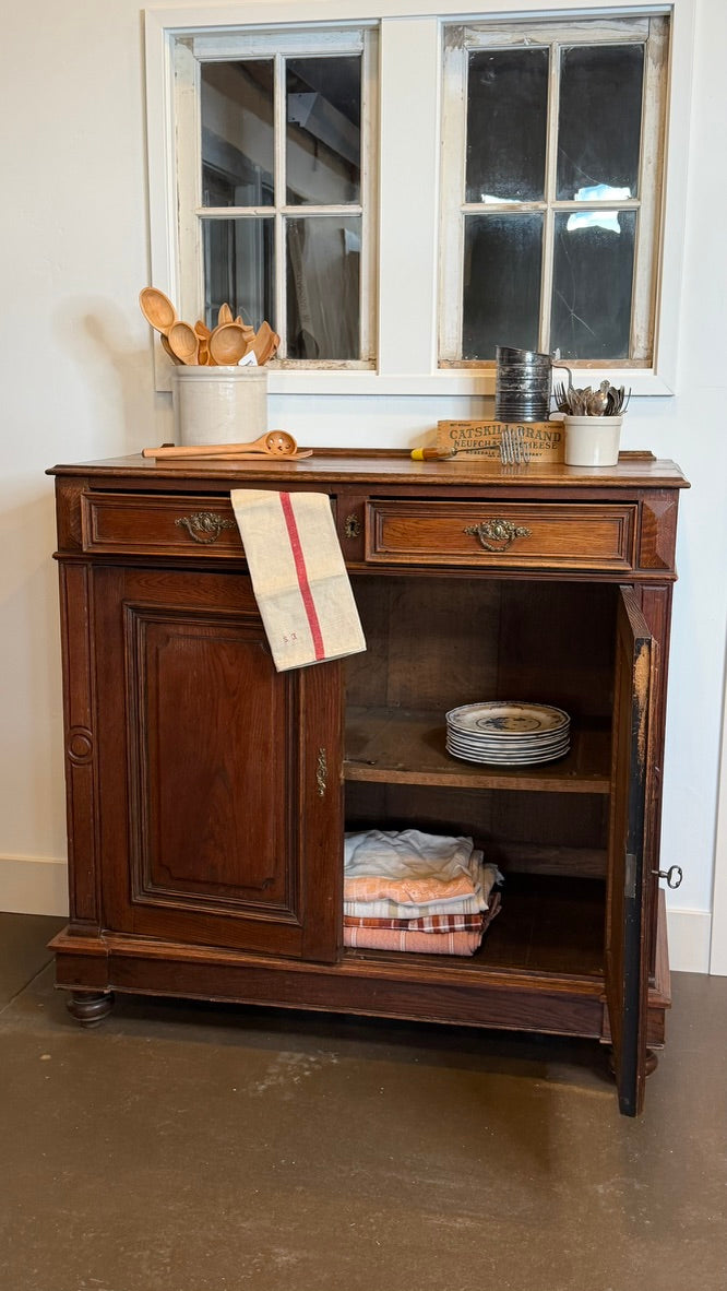 A dark brown wooden kitchen sideboard cabinet with an open door, styled with kitchen utensils, plates, and folded linens inside, set against a white wall with two windows.