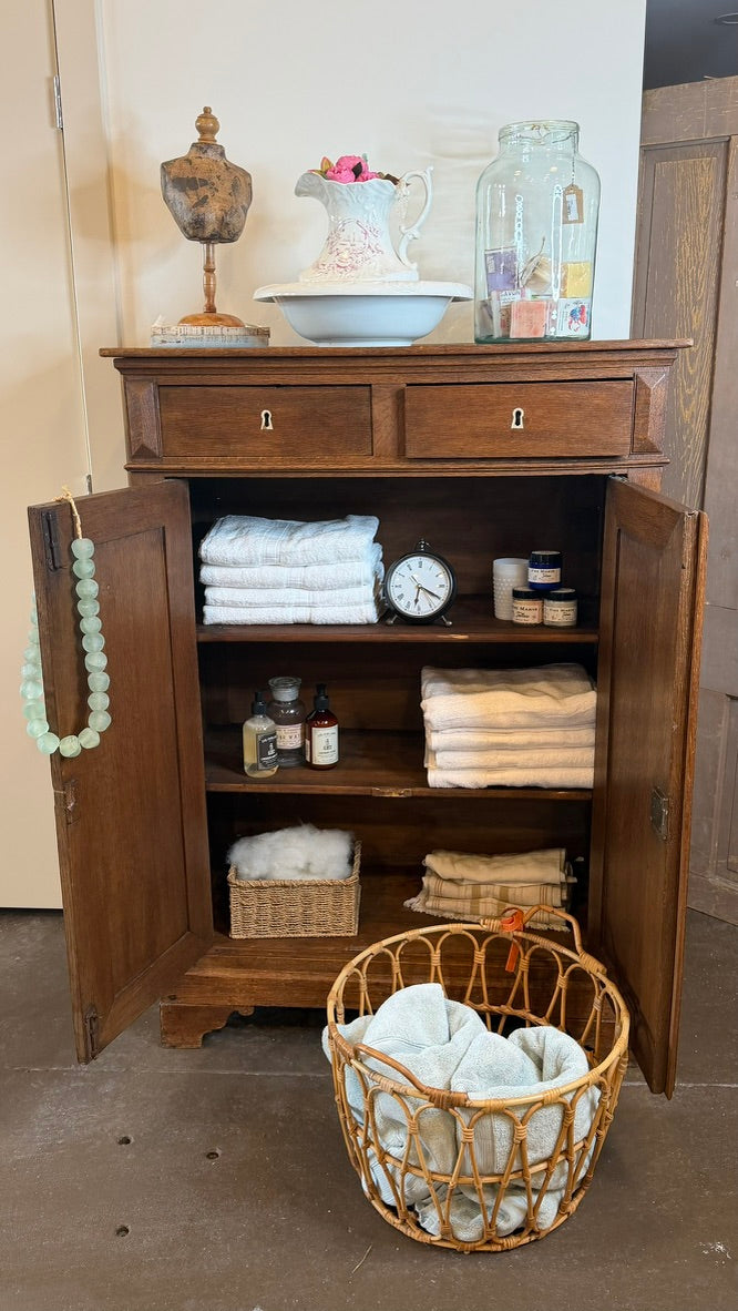 A rustic wooden bathroom cabinet with open doors, styled with folded towels, toiletries, and a decorative pitcher on top, with a wicker basket of folded linens in front on a concrete floor.