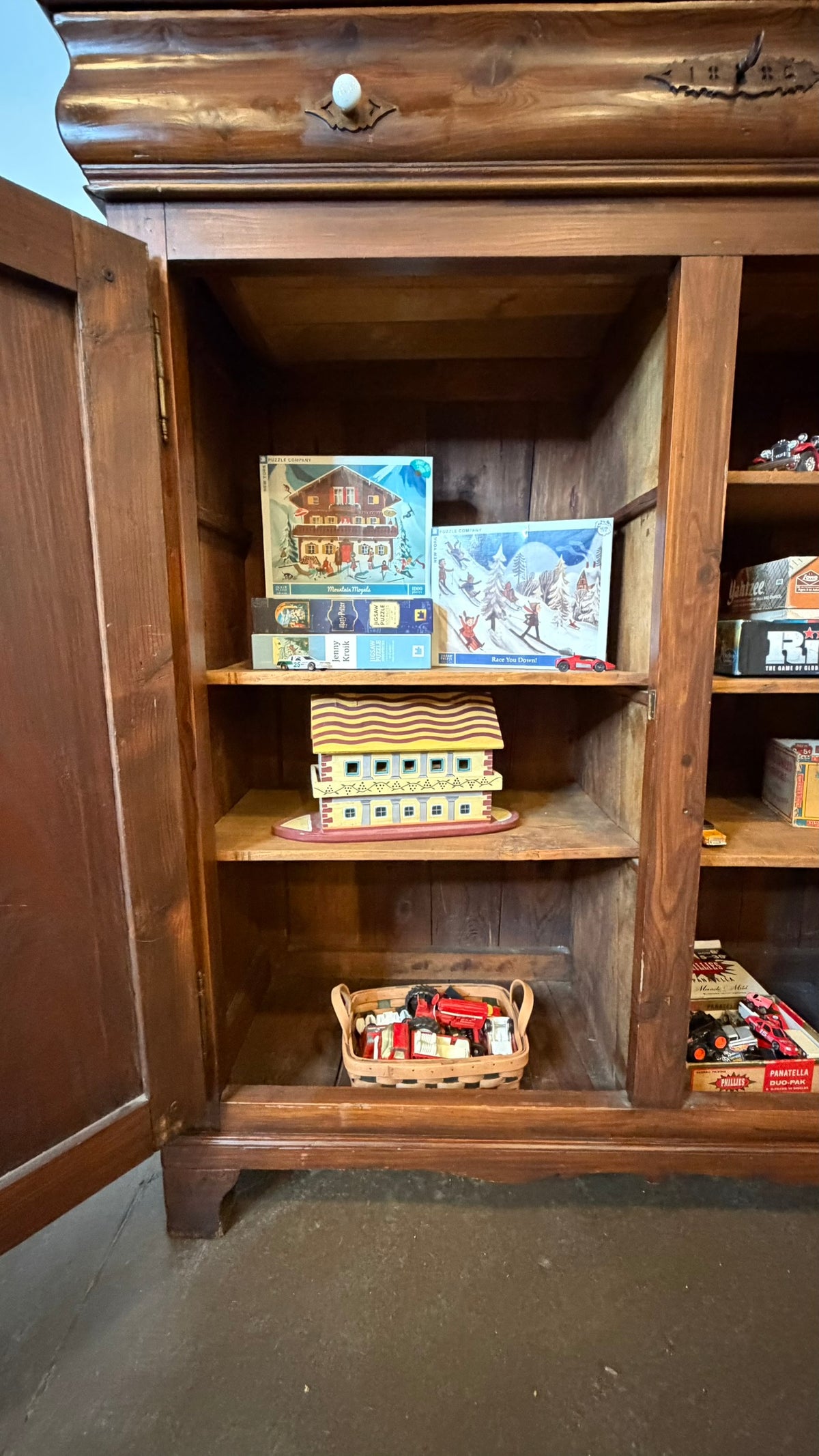 A rustic dark brown wooden cabinet with an open door, displaying shelves filled with various toy boxes, board games, and a small wicker basket on a concrete floor.