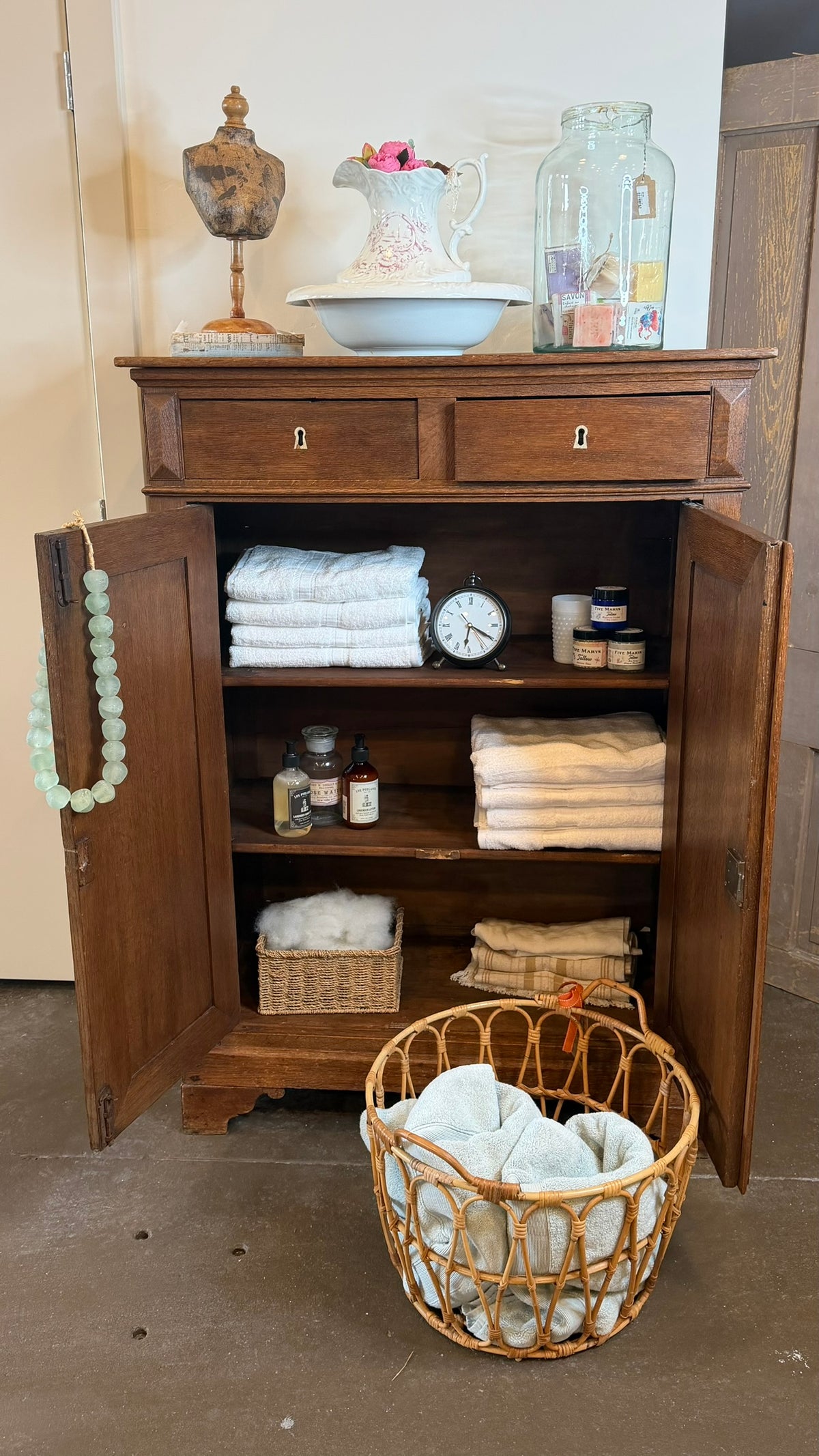 A rustic wooden linen cabinet with open doors, styled with folded white towels, small bottles, a clock, and decorative items on top, with a wicker basket of folded linens in front on a concrete floor.