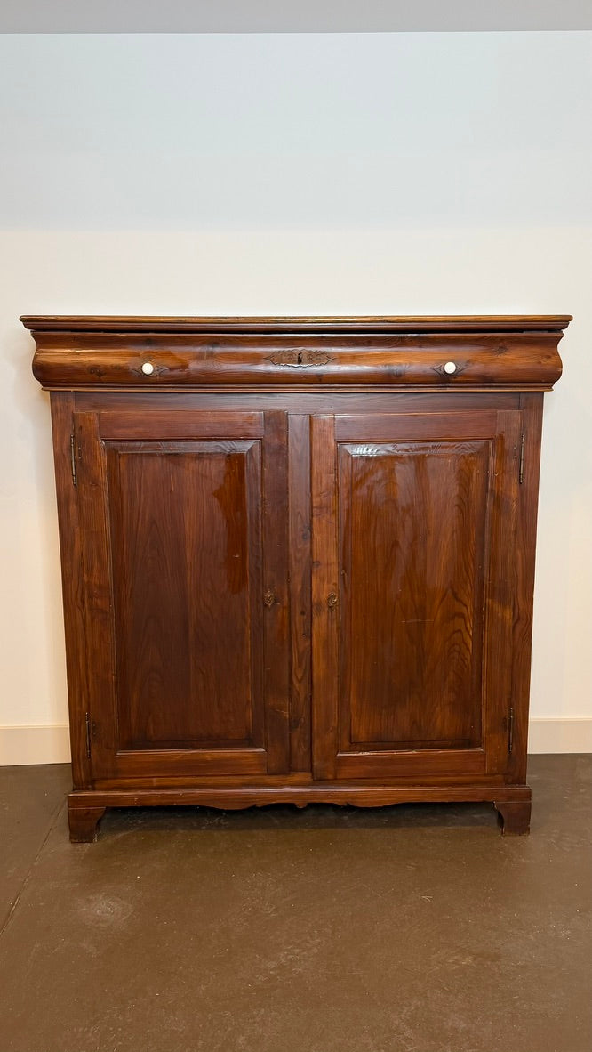 A dark brown wooden cabinet with two paneled doors and a decorative top, photographed against a plain light wall and brown floor.