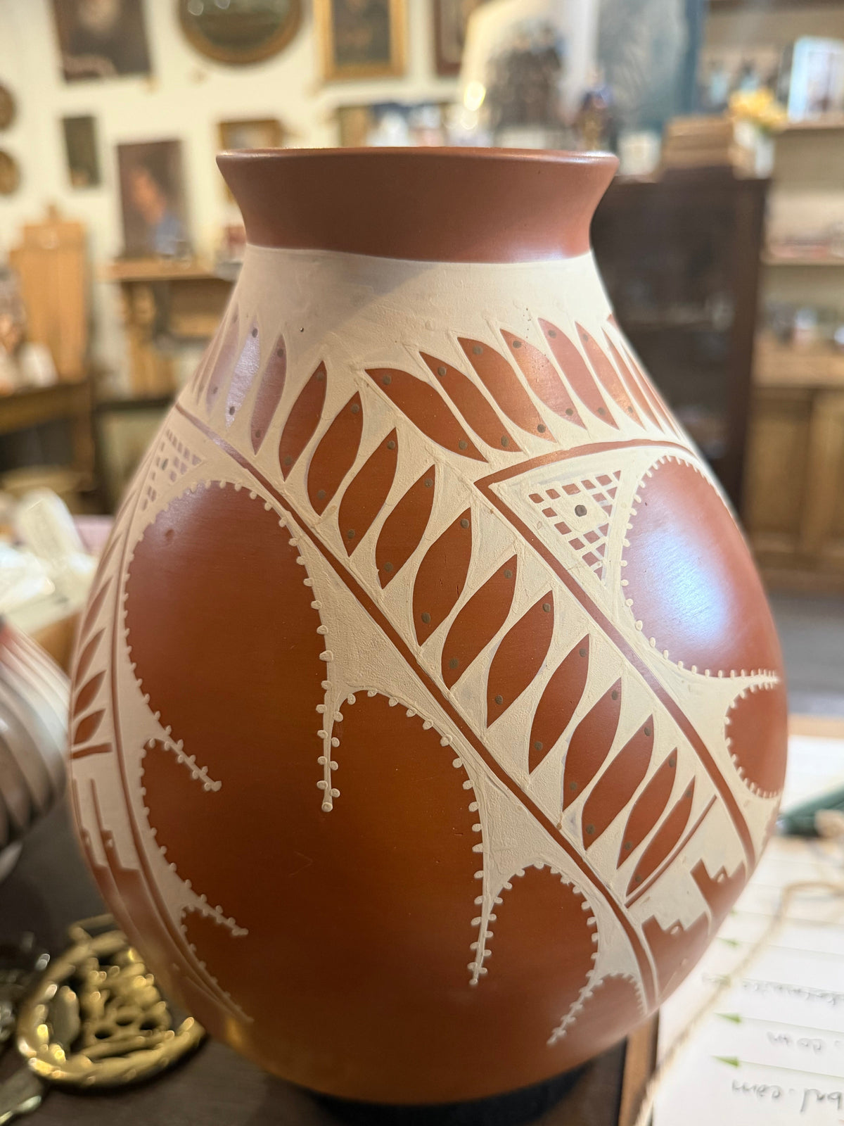 A terracotta clay vase with hand-painted geometric patterns in brown and cream, displayed on a table in what appears to be a pottery or home decor shop.