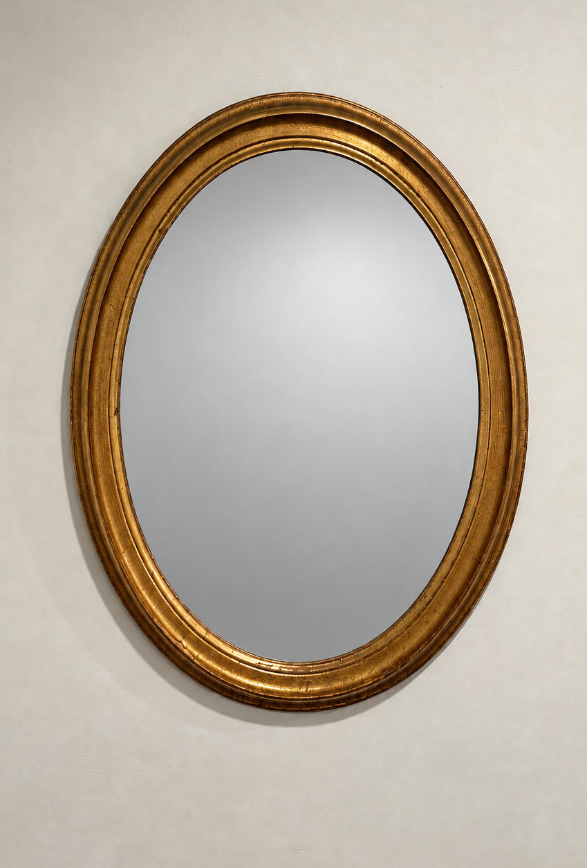An oval wall mirror with a thick, ornate gold frame, shown on a plain off-white wall. 