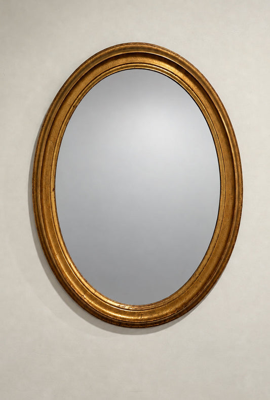 An oval wall mirror with a thick, ornate gold frame, shown on a plain off-white wall. 