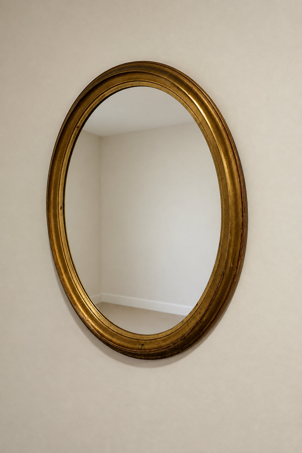 An oval wall mirror with a distressed gold frame, mounted on a plain off-white wall. The reflection in the mirror shows a green storage bin on a shelf below.