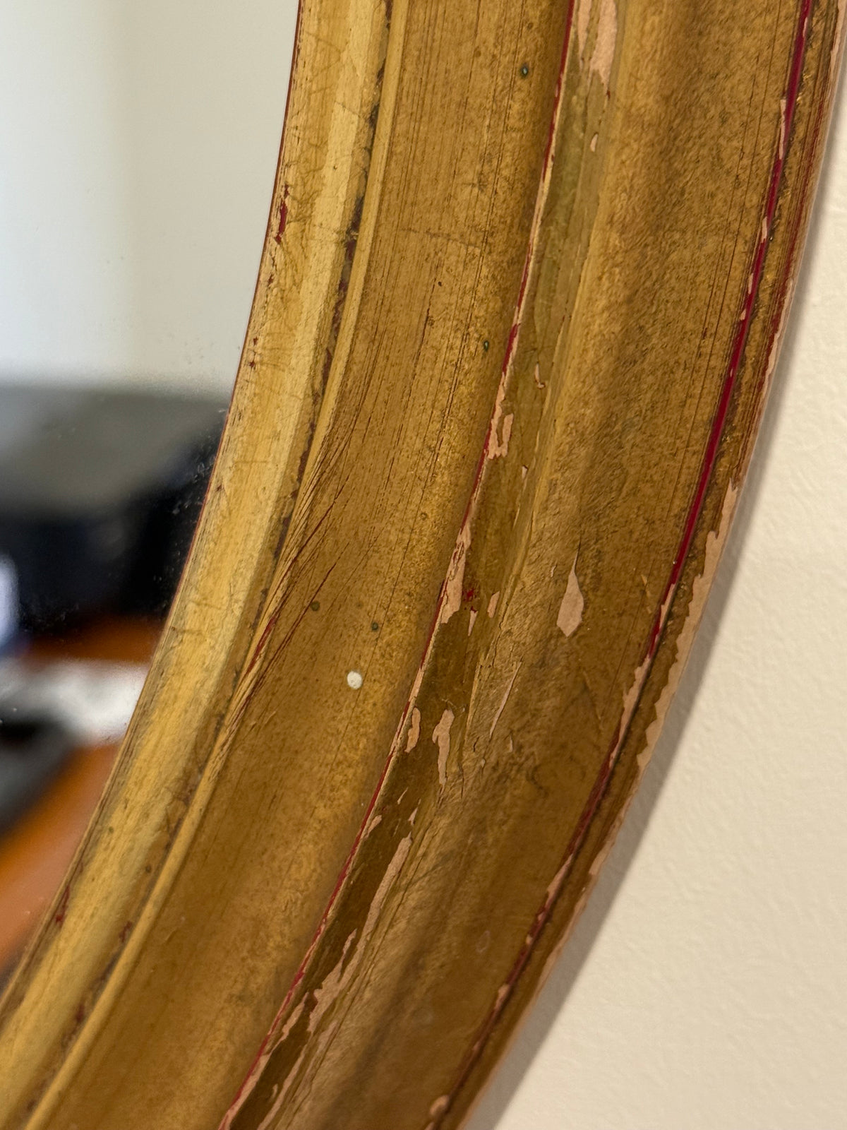 Close-up of a distressed gold mirror frame with peeling paint and visible red inner lining, set against a light wall.