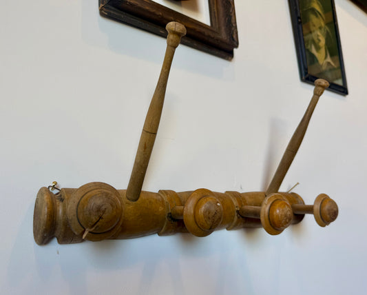 A vintage wooden wall-mounted coat rack with four pegs, shown hanging on a white wall with framed pictures partially visible in the background.