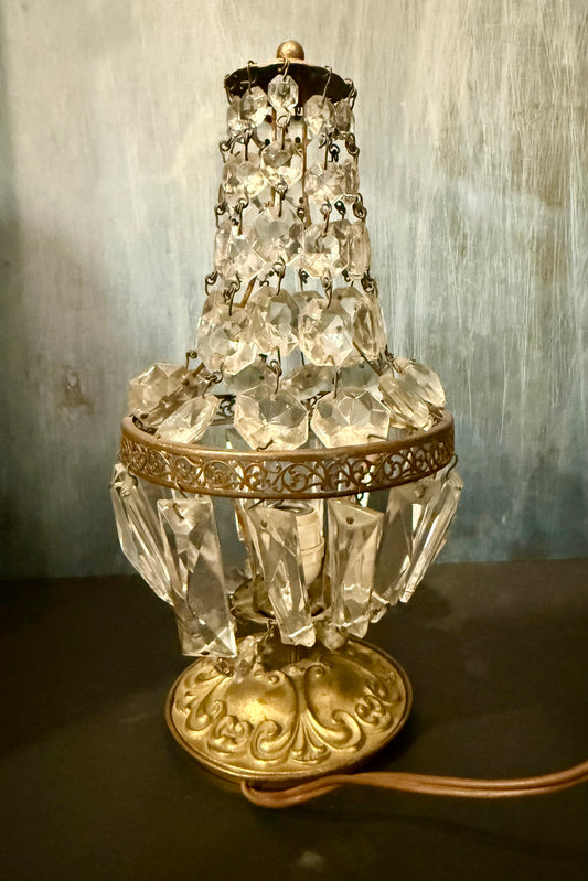 A vintage-style crystal and brass table lamp with clear faceted glass drops and ornate gold-toned metal detailing, shown on a dark surface against a textured gray wall.