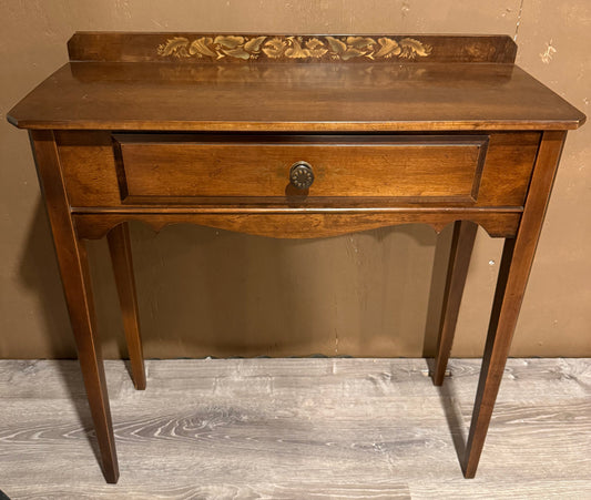 A small wooden console table with a single drawer, turned legs, and a decorative carved panel on the back of the top, shown against a textured tan wall and light wood floor.