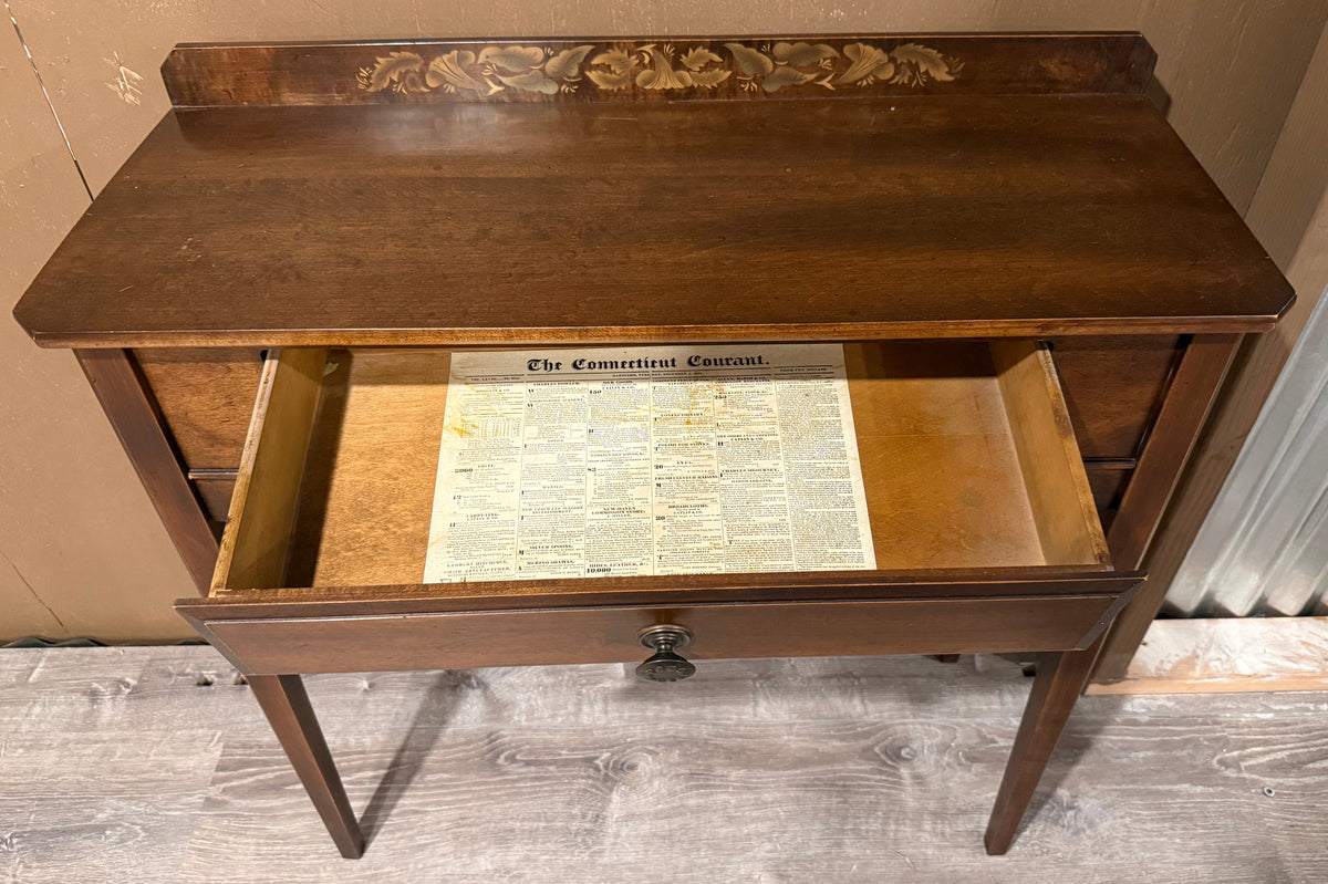 A vintage wooden writing desk with one drawer pulled open, showing a printed page inside. The desk has a dark brown wood finish, a decorative carved panel on the backboard, and a single round metal knob on the front drawer.