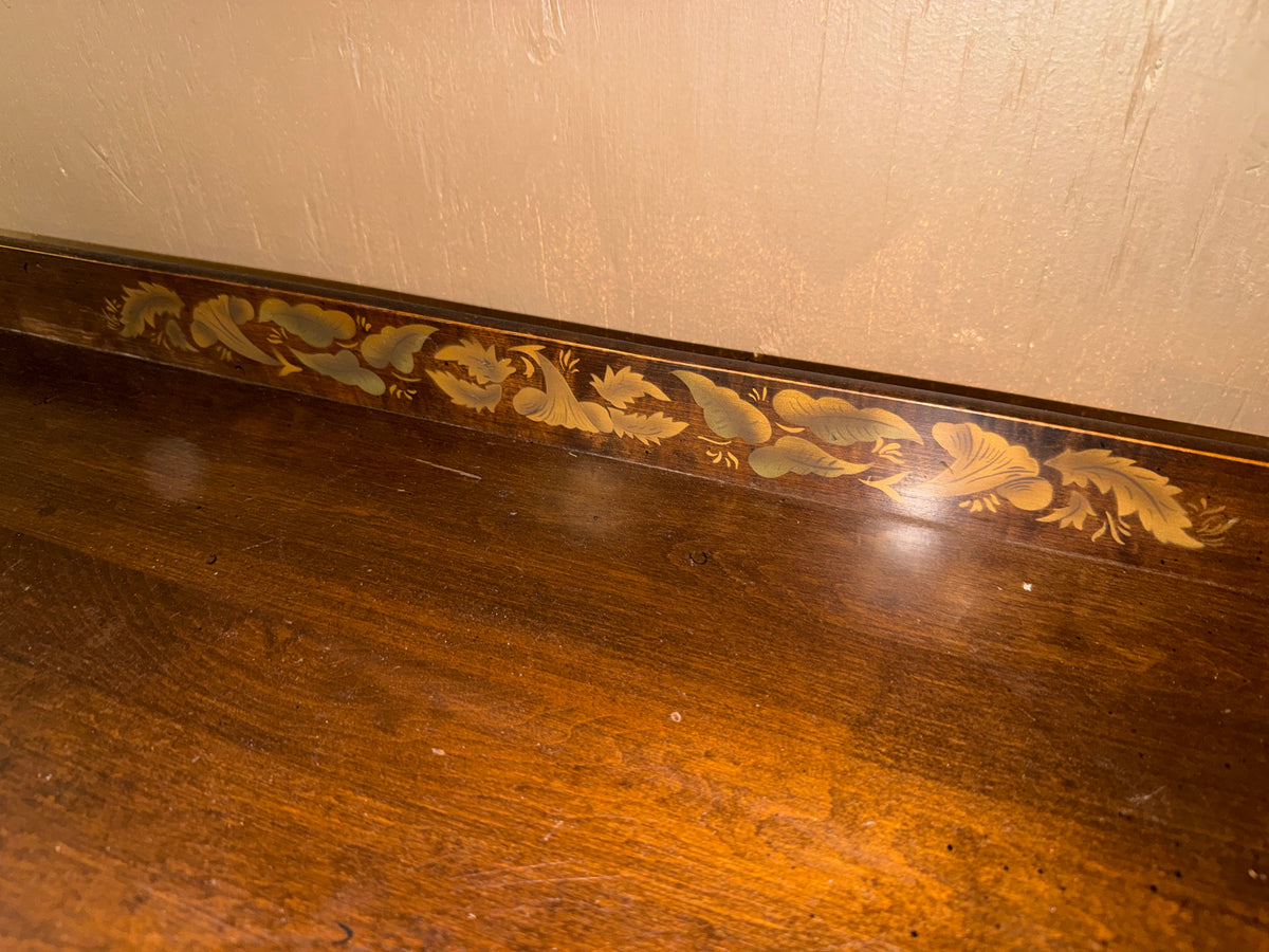 Close-up of a dark brown wooden surface with a decorative inlaid border featuring gold and light blue floral motifs, set against a beige wall.