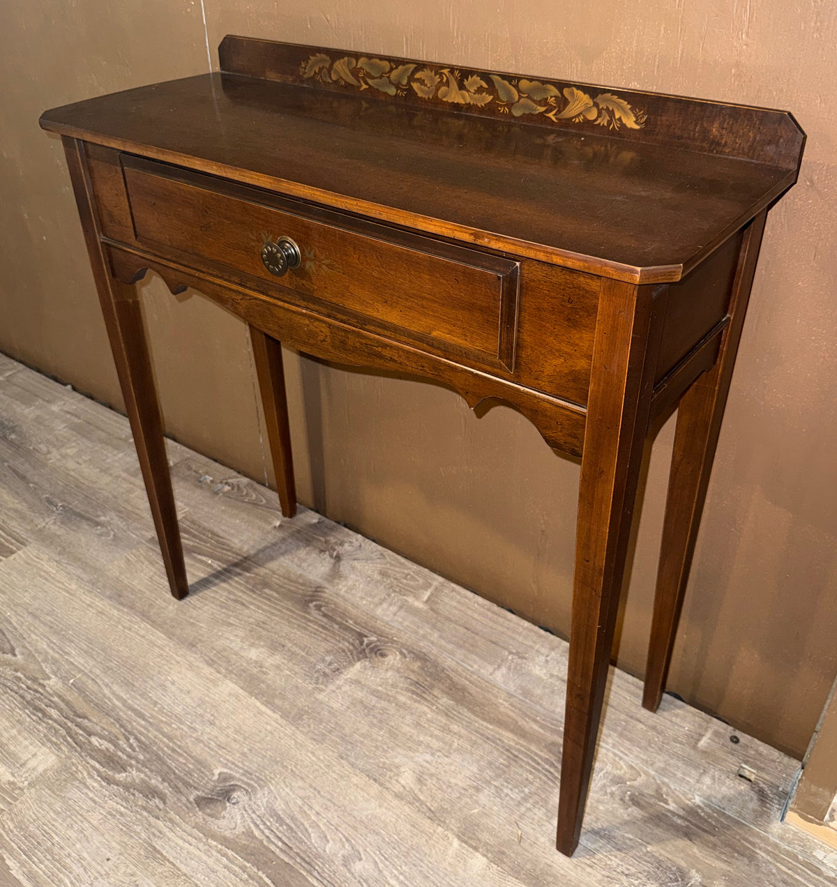 A small wooden console table with a single drawer, turned legs, and a decorative carved backrest, shown against a plain wall on a wood-look floor.