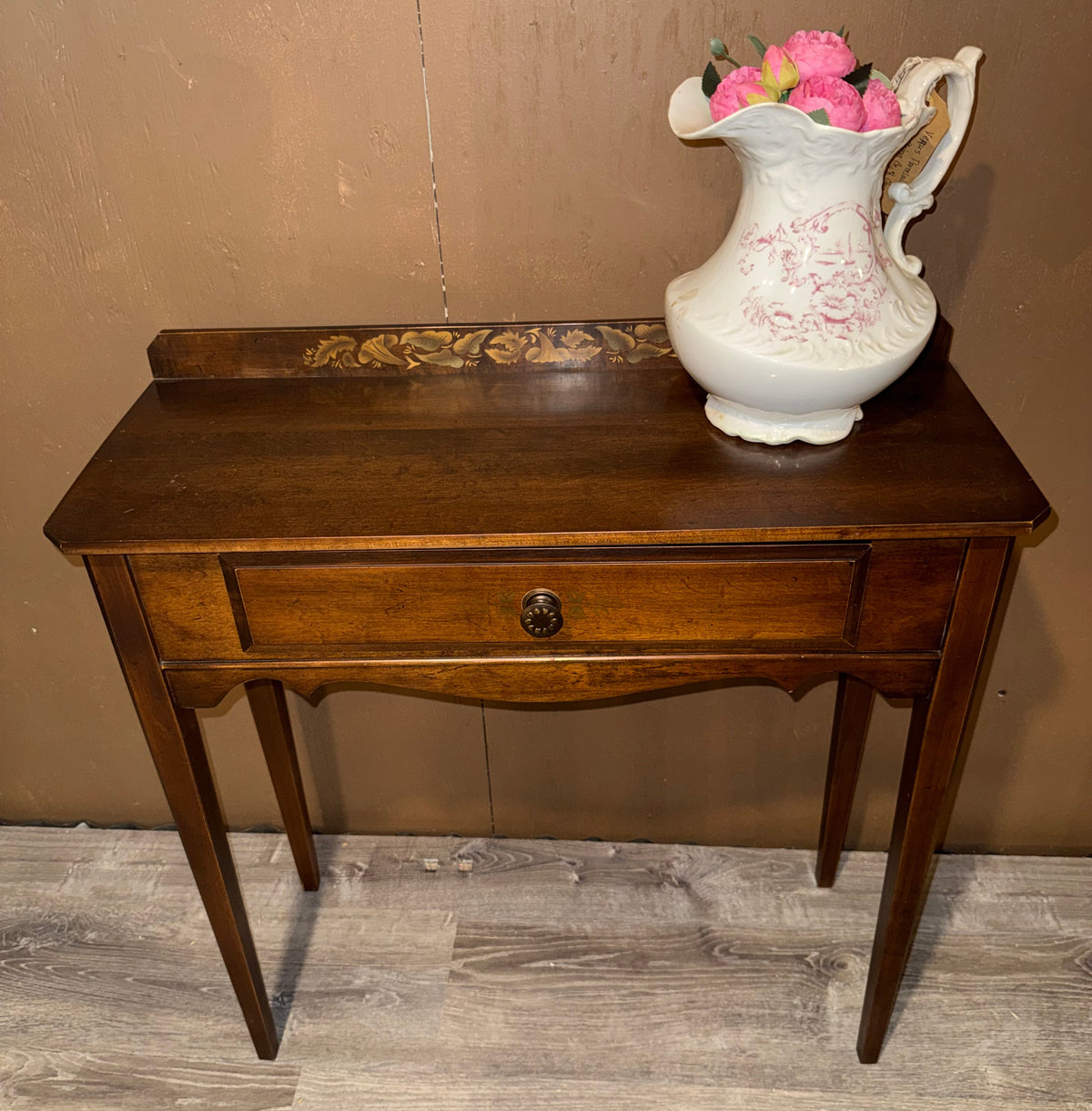 A dark brown wooden console table with a single drawer and curved apron, shown against a brown wall and gray wood-look floor. A white ceramic pitcher with pink floral decoration and pink flowers sits on the tabletop.