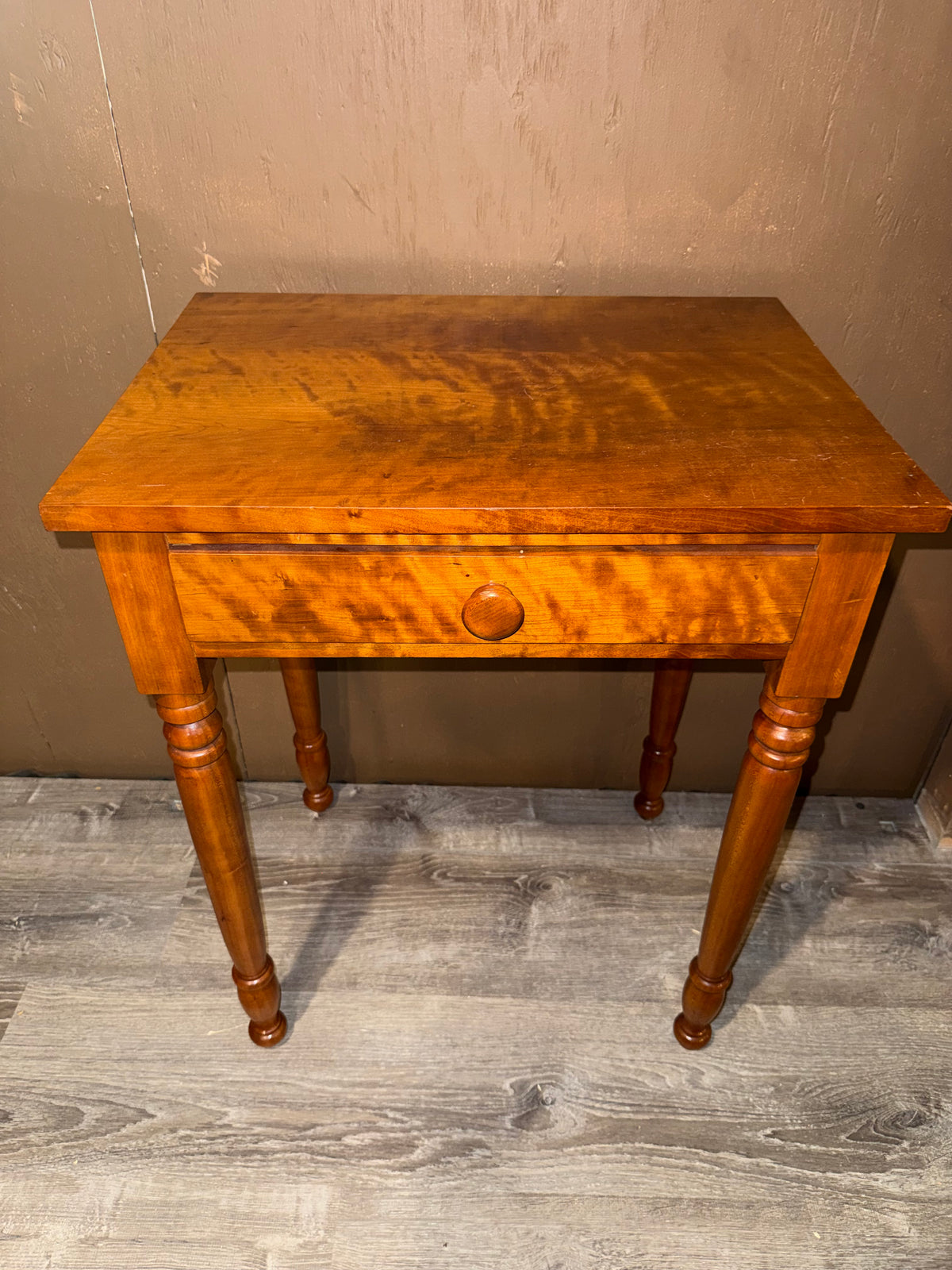 A small wooden writing desk with a single drawer and turned legs, shown against a textured brown wall and gray wood-look floor.
