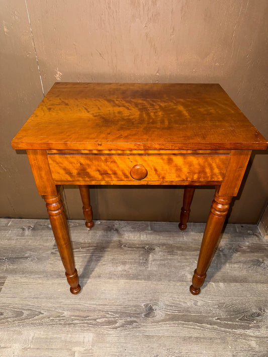 A small wooden writing desk with a single drawer and turned legs, shown against a textured brown wall and gray wood-look floor.